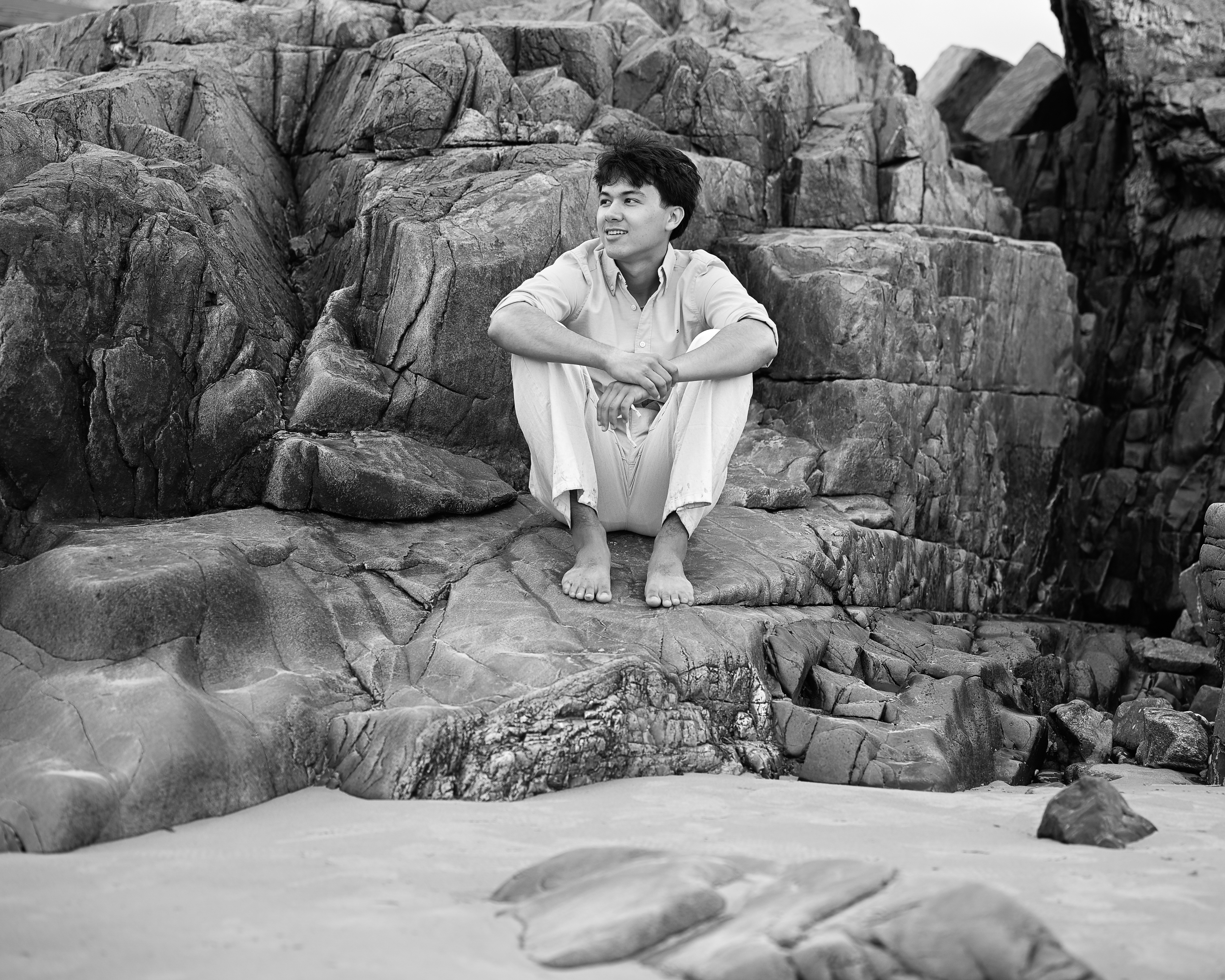 Black and white senior portrait seated on the rocky shoreline at Fort Foster