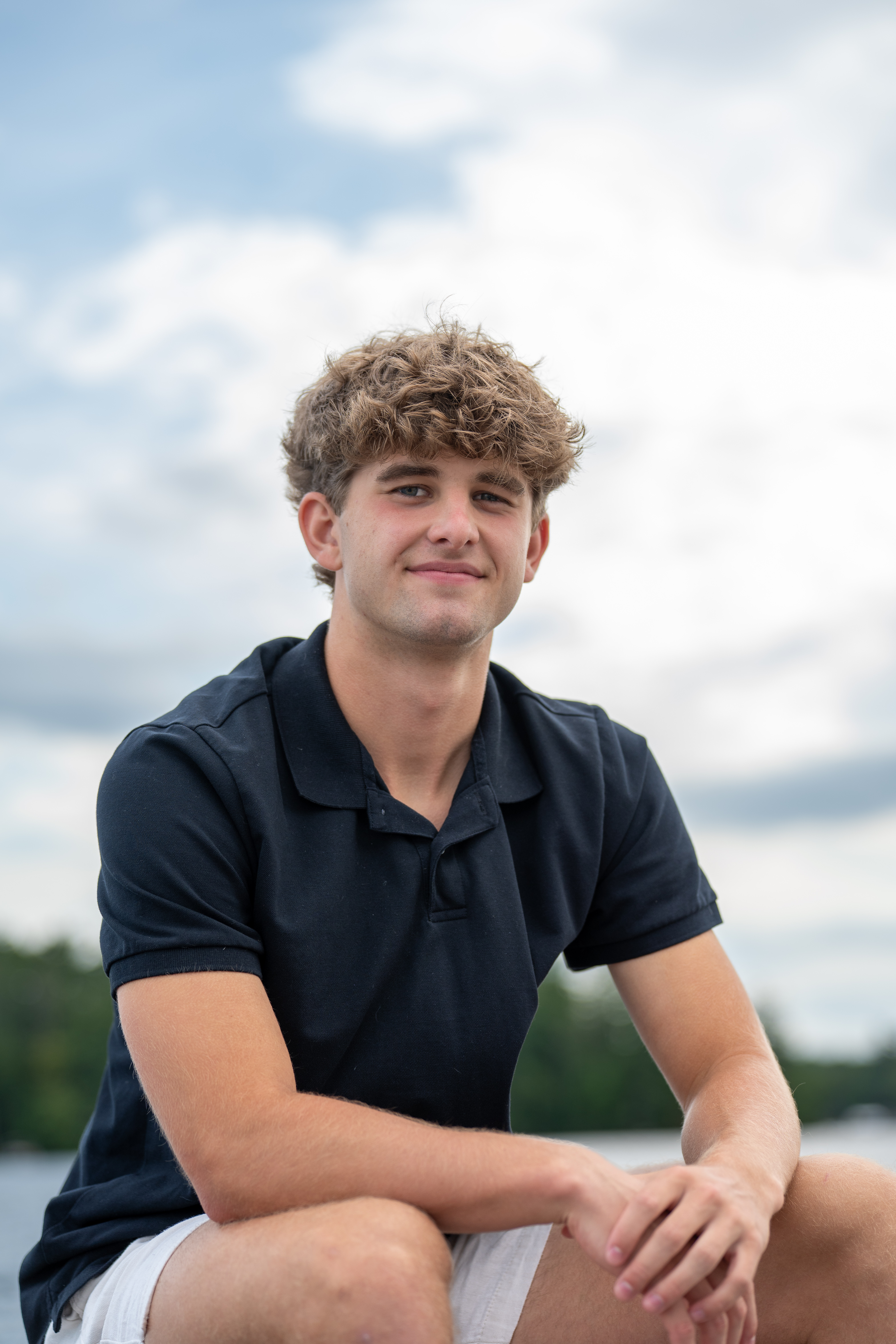 High school senior in a navy polo sitting by a lake under a cloudy sky, New Hampshire senior portrait