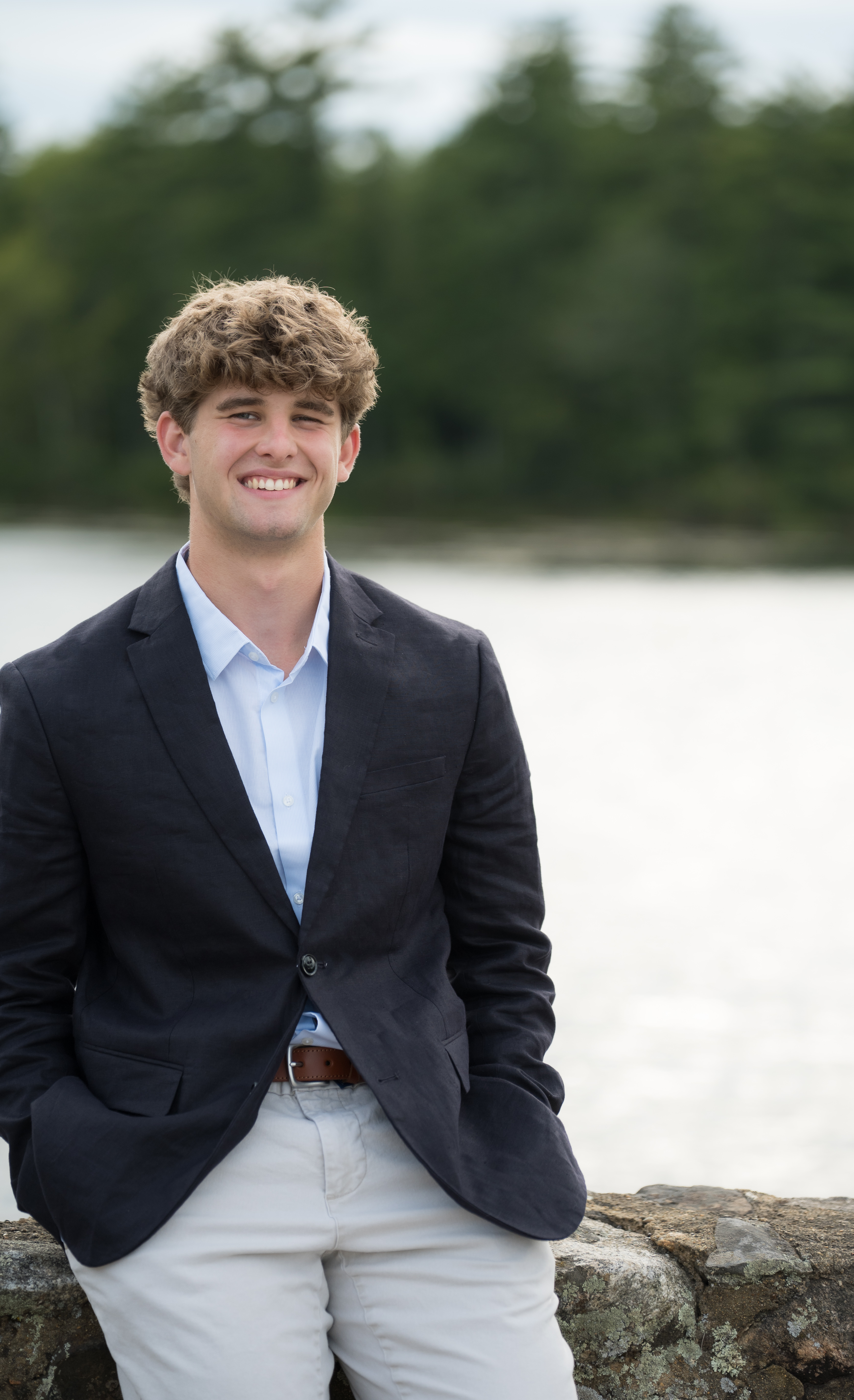 Smiling high school senior in a navy blazer leaning on a stone wall by a lake, New Hampshire senior portrait